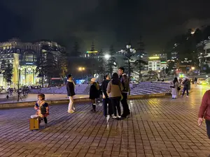 Quang cảnh Sapa về đêm tại khu vực quảng trường trung tâm. Nổi bật là tòa nhà Sun Plaza với kiến trúc châu Âu và đồng hồ lớn, đây cũng là lối vào Ga Sapa. Nhiều người dân và du khách mặc áo ấm đang tụ tập và đi dạo trên sân gạch. Ở phía trước, một em bé gái trong trang phục dân tộc đang đứng cạnh một chiếc hộp nhỏ.。（日本語訳）。中央広場エリアのサパの夜景。ヨーロッパ建築と大きな時計が特徴的なサンプラザビルが際立っており、ここはサパ駅の入り口でもある。暖かい服を着た多くの地元の人々や観光客が、レンガ敷きの広場に集まったり散策したりしている。手前には、民族衣装を着た一人の少女が小さな箱の隣に立っている。