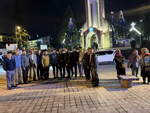 Một nhóm đông người, chủ yếu là đàn ông, đang đứng chụp ảnh tập thể vào ban đêm trước một nhà thờ đá. Nhà thờ được trang trí lộng lẫy bằng đèn neon nhiều màu sắc và bên cạnh có những cây thông Noel cũng được thắp sáng. Ở phía bên phải, có một vài người phụ nữ, trong đó có người mặc trang phục dân tộc thiểu số, đứng gần một chiếc ghế nhỏ bày bán các món đồ trang sức.。（日本語訳）。主に男性からなる大勢のグループが、夜、石造りの教会の前で集合写真を撮っています。教会は色とりどりのネオンで豪華に飾られており、その隣にはライトアップされたクリスマスツリーがあります。右側には、少数民族の衣装を着た人を含む数人の女性が、アクセサリーを並べて売っている小さな椅子の近くに立っています。