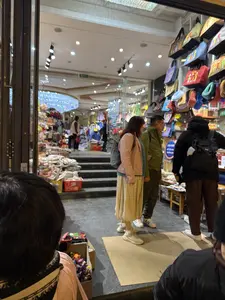 Bên trong một cửa hàng bán đồ lưu niệm, có nhiều khách hàng đang lựa chọn sản phẩm. Ở phía trước, một nhóm người đang đứng xem hàng. Một người phụ nữ mặc áo khoác màu hồng, váy dài màu be, đứng cạnh một người đàn ông mặc áo khoác màu xanh. Một người khác mặc đồ đen đang xem các món đồ được bày bán. Cửa hàng được chiếu sáng rực rỡ, bên trong có nhiều quần áo và hàng hóa khác. Ở phía ngoài, trên tường treo rất nhiều túi xách bằng vải với đủ màu sắc.。（日本語訳）。お土産屋の中では、多くのお客さんが商品を選んでいます。手前では、一団の人々が立って商品を見ています。ピンク色のジャケットを着て、ベージュ色のロングスカートを履いた女性が、青いジャケットを着た男性の隣に立っています。もう一人の黒い服を着た人が、陳列されている品々を見ています。店内は明るく照らされており、中にはたくさんの服やその他の商品があります。外側の壁には、様々な色の布製ハンドバッグがたくさん掛けられています。