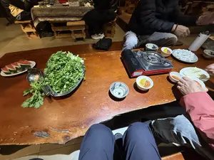 Một nhóm người đang quây quần bên một chiếc bàn gỗ lớn để ăn lẩu. Trên bàn có một bếp ga mini, một đĩa rau xanh lớn, một đĩa dưa hấu, một đĩa đậu phụ và một đĩa sủi cảo. Ngoài ra còn có nhiều bát nhỏ đựng nước chấm. Mọi người ngồi trên những chiếc ghế gỗ thấp trong một không gian ấm cúng.。（日本語訳）。あるグループの人々が、鍋を食べるために大きな木製のテーブルを囲んでいます。テーブルの上には、ミニカセットコンロ、青菜の大皿、スイカの皿、豆腐の皿、そして水餃子の皿があります。その他にも、つけダレを入れた小鉢がたくさんあります。皆は、居心地の良い空間で、低い木製の椅子に座っています。