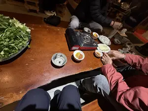 Bức ảnh chụp cảnh một nhóm người đang quây quần bên một chiếc bàn gỗ để chuẩn bị ăn lẩu. Trên bàn có một bếp ga mini, một đĩa rau xanh lớn, một đĩa đậu phụ thái miếng, một đĩa chả viên và các bát nhỏ đựng nước chấm. Mọi người đang ngồi trên ghế thấp hoặc ngồi bệt quanh bàn, chuẩn bị cho bữa ăn trong một không khí ấm cúng.。（日本語訳）。木製のテーブルを囲んで鍋の準備をしている人々のグループの写真です。テーブルの上には、ミニガスコンロ、大皿の青野菜、スライスされた豆腐の皿、つみれの皿、そしてつけダレが入った小鉢があります。皆は低い椅子に座ったり、床に座ったりしてテーブルを囲み、温かい雰囲気の中で食事の準備をしています。