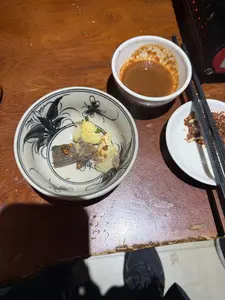 Trên một chiếc bàn gỗ, có một bát gốm sứ hoa văn đen trắng đựng vài miếng thức ăn trông giống như lòng luộc, có rắc thêm ớt bột và hành lá. Bên cạnh là một bát nước chấm màu nâu và một đĩa nhỏ đựng gia vị ớt bột cùng một đôi đũa đen.。（日本語訳）。隣には茶色のつけダレの碗と、唐辛子パウダーのスパイスが入った小皿、そして黒い箸一膳がある。