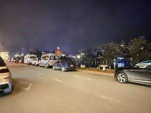 Cảnh một con đường vào ban đêm, có nhiều xe ô tô đang đỗ hai bên. Bên trái là một hàng xe, bao gồm xe van trắng, xe van bạc và một chiếc xe con màu xanh dương. Bên phải đường là một hồ nước, mặt hồ phản chiếu ánh đèn lung linh từ các tòa nhà và đèn trang trí ở phía xa. Bầu trời đêm tối và có vẻ như có sương mù hoặc khói.。（日本語訳）。夜の道の一場面で、両側に多くの自動車が駐車している。左側は車の列で、白いバン、シルバーのバン、そして一台の青い乗用車がある。道路の右側は湖で、湖面は遠くの建物や装飾灯からのきらめく光を反射している。