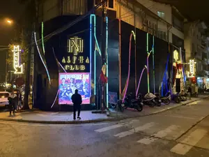 Bức ảnh chụp cảnh một câu lạc bộ đêm tên là "Alpha Club" vào ban đêm. Câu lạc bộ nằm ở góc một con phố, có mặt tiền màu đen được trang trí bằng nhiều dải đèn neon nhiều màu sắc. Có một tấm áp phích lớn được chiếu sáng quảng cáo cho một sự kiện "Independence Day", và một người đang đứng nhìn vào đó. Nhiều xe máy được đỗ trên vỉa hè bên cạnh câu lạc bộ. Một lá cờ Việt Nam cũng được treo gần lối vào.。（日本語訳）。「アルファクラブ」という名のナイトクラブを夜に撮影した写真です。クラブは街角にあり、黒い正面は色とりどりの多くのネオンサインで飾られています。「独立記念日」のイベントを宣伝する、ライトアップされた大きなポスターがあり、一人の人がそれを見て立っています。クラブの隣の歩道には多くのバイクが停められています。入り口の近くにはベトナム国旗も掲げられています。