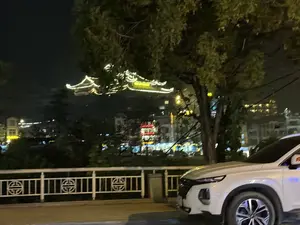 Bức ảnh chụp cảnh đêm tại một thành phố. Ở phía trước, một phần của chiếc ô tô màu trắng đang đỗ bên đường. Bên cạnh là lan can màu trắng có hoa văn cổ điển. Phía sau cây cối, một công trình kiến trúc kiểu Á Đông trên đồi được thắp sáng nổi bật với những đường viền ánh sáng vàng. Xa hơn là khung cảnh thành phố về đêm với những tòa nhà và ánh đèn nhiều màu sắc bị nhòe đi.。（日本語訳）。ある都市の夜景を撮影した写真です。手前には、道路脇に駐車している白い車の一部が見えます。その隣には、クラシックな模様の白い手すりがあります。木々の後ろには、丘の上の東アジア風の建築物が金色の光の輪郭でライトアップされ、際立っています。さらに遠くには、建物や色とりどりの光がぼやけた、街の夜景が広がっています。