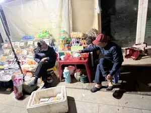 Vào ban đêm, tại một quán nước vỉa hè, một người phụ nữ đang ngồi trên chiếc ghế nhựa màu vàng. Bà mặc áo khoác tối màu và đeo khẩu trang. Đối diện bà, một người đàn ông đội mũ lưỡi trai màu đỏ, mặc bộ đồ thể thao, đang ngồi trên ghế xanh và rót trà từ ấm trắng vào cốc. Giữa họ là chiếc bàn nhựa màu đỏ. Xung quanh có nhiều phích nước và ấm đun. Phía sau họ là một quầy hàng tạm bợ bán nhiều loại đồ ăn vặt đóng gói như ô mai, bánh kẹo và nước giải khát. Dưới đất có một thùng xốp trắng đựng tiền.。（日本語訳）。夜、とある道端の飲み物屋台で、一人の女性が黄色いプラスチックの椅子に座っている。彼女は暗い色のジャケットを着て、マスクを着けている。彼女の向かいには、赤い野球帽をかぶり、スポーツウェアを着た男性が緑の椅子に座り、白い急須からコップにお茶を注いでいる。彼らの間には赤いプラスチックのテーブルがある。周りには多くの魔法瓶や湯沸かし器がある。彼らの後ろには、オーマイ（ドライフルーツの砂糖漬け）やお菓子、清涼飲料水など、多くの種類の袋入りスナックを売る簡易的な売店がある。地面にはお金を入れた白い発泡スチロールの箱がある。