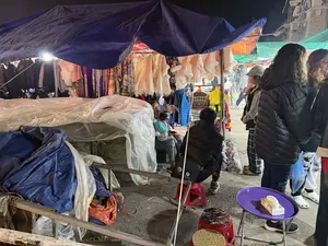 Cảnh một gian hàng quần áo tại một khu chợ đêm. Gian hàng được dựng tạm bằng khung kim loại và che bằng tấm bạt lớn màu xanh. Nhiều loại quần áo mùa đông được treo trên giá, bao gồm áo khoác phao, áo len trắng có cổ lông và khăn choàng với nhiều hoa văn khác nhau. Một số khách hàng đang đứng xem và lựa chọn đồ. Một người phụ nữ, có thể là chủ cửa hàng, đang ngồi bế một đứa trẻ nhỏ. Một người khác ngồi trên chiếc ghế đẩu nhựa màu đỏ. Phía trước có một chiếc bàn nhỏ màu tím trên đó có một hộp khăn giấy. Không khí chợ đêm trông khá tấp nập.。（日本語訳）。あるナイトマーケットの衣料品店の光景。その店は金属フレームで仮設され、大きな青い防水シートで覆われている。ダウンジャケット、ファーの襟が付いた白いセーター、様々な柄のショールなど、多くの種類の冬服がラックに掛けられている。何人かの客が立って商品を見て、選んでいる。店主と思われる一人の女性が、小さな子供を抱いて座っている。別の人は赤いプラスチック製のスツールに座っている。