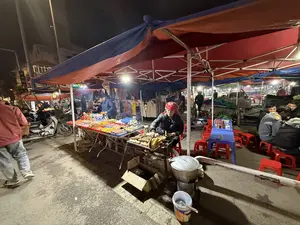 Khung cảnh một khu chợ đêm ở Việt Nam, tấp nập và nhộn nhịp. Dưới những mái lều bạt màu đỏ và xanh, các gian hàng được thắp sáng bằng đèn điện. Ở trung tâm bức ảnh, một người phụ nữ đội khăn đỏ, mặc áo khoác da đen đang ngồi bán hàng, tay đang chuẩn bị đồ ăn nướng như ngô, trứng và các loại xiên que. Bên cạnh là một quầy hàng khác bày bán nhiều loại xiên que màu sắc và nước ngọt. Một vài khách hàng đang lựa chọn đồ ăn. Ở phía sau, một nhóm người đang ngồi ăn uống trên những chiếc ghế nhựa màu đỏ và xanh dương. Phía xa có các gian hàng bán quần áo và một biển hiệu sáng đèn có chữ "BIDV". Một chiếc xe máy được dựng gần đó. Dưới đất có một chiếc nồi nhôm lớn và một thùng sơn hiệu Dulux.。（日本語訳）。ベトナムのあるナイトマーケットの光景、混雑して賑やかだ。赤と青の防水シートの屋根の下、各屋台は電灯で照らされている。写真の中央では、赤いスカーフをかぶり、黒い革のジャケットを着た女性が座って商品を売っており、手はトウモロコシ、卵、各種串焼きなどの焼き物を準備している。隣には、色とりどりの串焼きやソフトドリンクを並べた別の屋台がある。何人かの客が食べ物を選んでいる。後方では、赤と青のプラスチックの椅子に座って飲食している人々のグループがいる。遠くには衣料品を売る屋台と、「BIDV」という文字が書かれた光る看板がある。近くにバイクが一台停められている。地面には大きなアルミ鍋とDuluxブランドのペンキ缶がある。