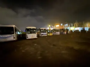 Bức ảnh chụp cảnh ban đêm, bị mờ và nhòe do chuyển động. Trong ảnh có một số chiếc xe buýt du lịch màu trắng đang đỗ trên một bãi đất. Bầu trời tối và có mây. Ở phía sau, có thể thấy ánh đèn từ các tòa nhà bị nhòe đi, tạo thành các vệt sáng màu vàng và trắng.。（日本語訳）。夜景を撮影した写真で、動きによりぼやけて不鮮明です。空は暗く、雲があります。背景では、建物からの光がにじみ、黄色と白の光の筋を形成しているのが見えます。