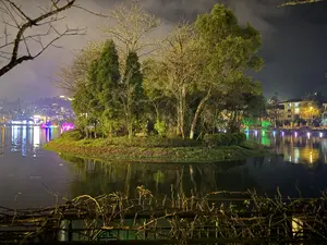 Cảnh đêm tại một hồ nước, với một hòn đảo nhỏ ở trung tâm. Trên đảo có nhiều loại cây, một số có lá xanh tươi, một số khác thì khẳng khiu, trụi lá. Ánh đèn chiếu sáng hòn đảo và cây cối. Phía sau hòn đảo là một thị trấn ven hồ với nhiều tòa nhà sáng đèn. Ánh đèn từ các tòa nhà và đèn trang trí nhiều màu sắc trên mặt hồ phản chiếu xuống mặt nước tĩnh lặng, tạo nên một khung cảnh lung linh. Bầu trời đêm u ám và có mây. Ở phía trước, có thể thấy các cành cây khô của một hàng rào hoặc bụi cây.。（日本語訳）。ある湖の夜景、中央に小さな島がある。島には多くの種類の木があり、青々とした葉を持つものもあれば、葉が落ちて骨ばったものもある。照明が島と木々を照らしている。島の向こうには、多くの建物が明かりを灯す湖畔の町がある。建物からの光と湖上の色とりどりの装飾灯が静かな水面に反射し、きらびやかな光景を作り出している。夜空は暗く、雲がかかっている。