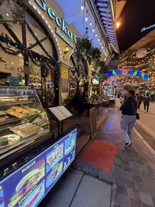 Khung cảnh một con phố tấp nập về đêm. Ở phía trước là nhà hàng Gecko với mặt tiền màu vàng, được trang trí đèn và dây kim tuyến. Bên ngoài có một tủ kính trưng bày bánh ngọt, một giá để thực đơn và bàn ghế cho khách ngồi. Một vài thực khách đang ngồi tại bàn. Trên vỉa hè, người qua lại đông đúc. Phía xa là một cổng chào lớn bằng đèn neon rực rỡ và các tòa nhà được thắp sáng bởi nhiều đèn lồng, tạo nên không khí sôi động của một khu phố du lịch.。（日本語訳）。夜の賑やかな通りの光景。手前には黄色いファサードのゲッコーレストランがあり、電飾とキラキラしたモールで飾られている。外にはケーキを陳列するガラスケース、メニューを置く台、そして客が座るためのテーブルと椅子がある。何人かの客がテーブルに座っている。歩道では、人々が賑やかに行き交っている。遠くには、色鮮やかなネオンでできた大きな歓迎ゲートがあり、建物は多くの提灯で照らされ、観光地の活気ある雰囲気を醸し出している。