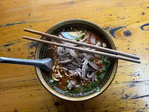 Một bát mì nóng hổi đặt trên mặt bàn gỗ màu vàng. Bát sứ có viền trang trí hoa văn màu đen. Bên trong bát là nước dùng màu nâu đỏ, sợi mì, rất nhiều thịt thái mỏng, nửa quả trứng luộc, và rắc nhiều hành lá cùng rau thơm. Một đôi đũa gỗ được đặt ngang trên miệng bát và một chiếc thìa inox nằm ở bên trái.。（日本語訳）。陶器の器には、縁に黒い模様の装飾がある。器の中には、赤褐色のスープ、麺、たくさんの薄切り肉、半分のゆで卵があり、たくさんのネギと香草が振りかけられている。