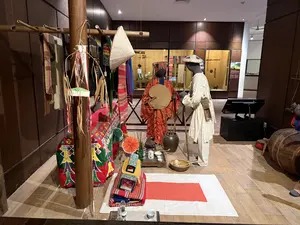 画像には、判読できる文字情報はありません。博物館の展示室で、民族衣装を着用した複数のマネキンが展示されています。木製の構造物には様々な布が掛けられ、床には儀式用具と思われる品々が並べられています
