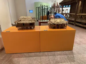 ベトナム民族学博物館の展示室で、オレンジ色の台座の上に藁葺き屋根の高床式住居の模型が2つ展示されている。画像内には判読可能な文字は写っていない