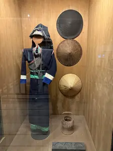 [ガラスケース内に、フード付きの青い民族衣装を着たマネキン、壁に掛けられた3つの笠、床に置かれた籠と模様入りの物体が展示されている