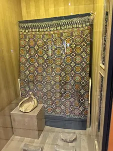 [Một tấm vải thổ cẩm với các hoa văn hình học được trưng bày trong tủ kính. Phía trước có một chiếc mũ hoặc giỏ và một vài vật thể khác.（幾何学模様が施された錦織の布がガラスケースの中に展示されている