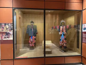 ガラスケースの中に、2体のマネキンが民族衣装を着て展示されている。左のマネキンは暗い色の上着とズボン、右のマネキンは赤を基調とした装飾的な衣装と頭飾りを身に着けている。ケースの両脇には説明パネルがあるが、文字は不鮮明で判読できない