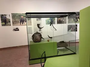 博物館の展示室。ガラスケースの中に、編みかご、動物の角で作られた道具などが展示されている。背景の壁には複数の写真が飾られているが、展示物の説明書きと思われるパネルの文字は不鮮明で判読できない
