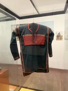 ガラスケースの中に展示されている、黒い長袖の上着。胸元には赤とオレンジの水平な織り模様の装飾があり、肩や袖にも模様が施されている