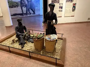 黒い服を着た2体のマネキンが、商いを営む様子を再現した展示物。座っている男性は履物のようなものを作り、立っている女性はカゴに入った商品を前にしている。背景には文字や写真を含む展示パネルがある