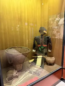 ガラスケースの中に、民族衣装を着たマネキン、編みかご、布製品などの民具が展示されている。奥にはモノクロ写真が掛けられている。手前の赤いラベルに文字があるが、不鮮明で判読できない