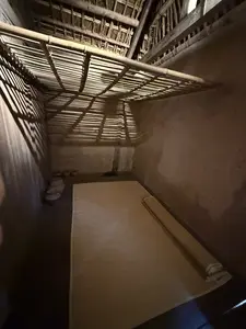 土壁の部屋の内部。床には敷物が置かれ、竹製の棚がある。天井は木と竹で組まれている