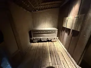 Bên trong một căn phòng nhỏ, tối, được tái hiện theo phong cách cổ xưa. Sàn nhà được làm bằng một tấm chõng tre lớn. Các bức tường được làm bằng đất và ván gỗ. Mái nhà lợp bằng lá với các thanh tre và gỗ lộ ra ngoài. Ở cuối phòng, dưới ánh đèn vàng, là một chiếc rương gỗ lớn, cũ kỹ với các chi tiết chạm khắc tinh xảo ở phần chân. Trên bức tường gỗ bên phải có treo ba tấm bảng ghi chú.。（日本語訳）。古風なスタイルで再現された、小さく暗い部屋の中。床は大きな竹製の寝台でできている。壁は土と木の板でできている。屋根は葉で葺かれており、竹と木の梁がむき出しになっている。部屋の奥、黄色い光の下に、脚部に精巧な彫刻が施された、大きくて古い木製の櫃がある。右側の木の壁には、3枚のメモ板が掛かっている。