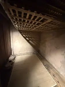 画像には文字がありません。暗い独房のような部屋が写っており、左奥の隅に便器のようなもの、床にござ、天井近くに木製の格子状の棚があります