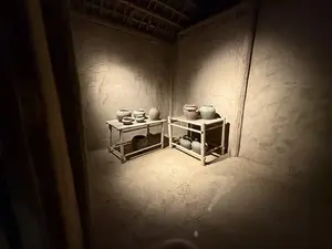 画像には文字は写っていません。薄暗い土壁のような部屋の中に、2つの木製の台が置かれ、その上に複数の陶器が展示されています。上からスポットライトで照らされています