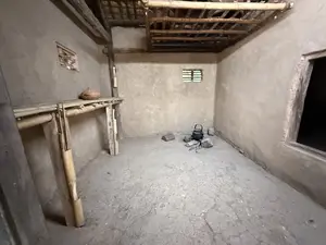 画像には、看板や説明文などの文字は一切含まれていません。土壁と竹の骨組みでできた部屋の内部。床は土間でひび割れている。壁際に竹製の棚があり、部屋の隅には釜が置かれた簡素な調理場がある