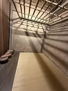 [画像には文字は写っていない。薄暗い部屋の内部。天井には木か竹でできた格子があり、その影が土壁と床に落ちている