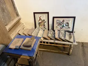 Trong ảnh là một góc trưng bày các dụng cụ làm tranh dân gian, có thể là tranh Đông Hồ. Dựa vào bức tường trắng là hai bức tranh đã được lồng khung kính. Một bức vẽ con cá chép màu vàng đỏ, và bức còn lại vẽ một gia đình gà. Trên các bức tranh có ghi chữ Hán Nôm. Phía trước, trên một chiếc ghế dài bằng gỗ, là một hàng các bản khắc gỗ (mộc bản) dùng để in tranh, với các hình khắc tinh xảo. Bên cạnh, trên một chiếc ghế đẩu thấp hơn được phủ vải xanh, có đặt thêm vài tấm ván gỗ và một chiếc chổi. Không gian mang dáng vẻ của một xưởng thủ công hoặc khu trưng bày về nghề làm tranh truyền thống.。（日本語訳）。写真には、民間絵画、おそらくはドンホー版画を制作するための道具を展示する一角が写っている。白い壁には、ガラスの額縁に入れられた2枚の絵が立てかけられている。一枚は赤みがかった黄色の鯉を描き、もう一枚は鶏の家族を描いている。絵の上には漢字・チュノムが書かれている。手前の木製の長い椅子の上には、絵を印刷するための、精巧な彫刻が施された木版（木版）が一列に並んでいる。この空間は、工芸工房、あるいは伝統的な絵画制作という職業に関する展示エリアの様相を呈している。