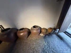 画像には、壁際に設置された石のような台の上に、大きさの異なる茶色の陶器の壺が複数並べられている。文字は写っていない