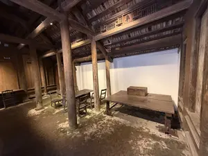 判読可能な文字情報は、この画像には含まれていません。画像は、木造建築の内部を写したものです。太い木の柱が床から天井を支え、奥には木製の机と椅子が置かれています。天井近くの梁には装飾的な彫刻が見られますが、文字として判読することはできません