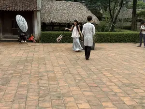 [画像には判読可能な文字は存在しない。煉瓦敷きの広場にて、伝統的な茅葺き屋根の家屋を背景に、アオザイのような衣装を着た男女が撮影を行っている