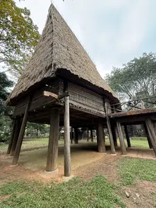 [画像には、茅葺きで急勾配の大きな屋根を持つ高床式の木造家屋が写っている。太い柱で支えられており、家屋に続く木製の通路がある