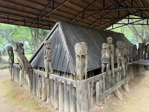 屋根付きの展示スペースに設置された、木製の柵と人型の木彫りの像で囲まれた木造の家]