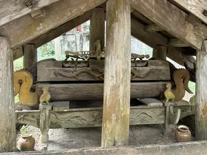 画像には判読可能な文字がありません。これは、木造の屋根の下に置かれた、龍や神話上の鳥などの彫刻が施された木製の棺のようなものです。(Không có chữ viết nào có thể đọc được trong hình ảnh. Đây là một vật thể bằng gỗ giống như quan tài, được chạm khắc hình rồng và các loài chim thần thoại, đặt dưới một mái che bằng gỗ.)