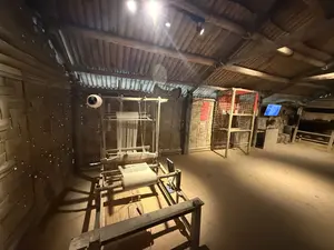 Khung cảnh trưng bày trong nhà với một khung dệt bằng gỗ. Tường và trần được lợp bằng tôn lượn sóng, phía sau có các sản phẩm làm từ tre. Không có chữ viết nào có thể đọc được.（木製の機織り機が置かれた室内の展示風景。壁と天井は波板で覆われ、奥には竹製品が見えます。判読できる文字はありません