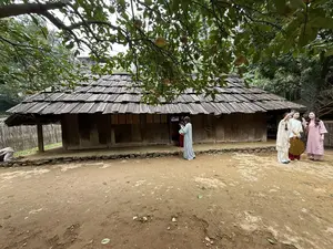 ベトナム民族学博物館の敷地内に展示されている、木製の板で葺かれた屋根を持つ伝統家屋。家の前には、伝統衣装を着用した数人の人物が立っている]