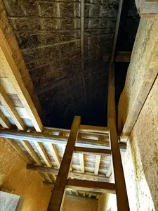 画像には文字がありません。土壁の家屋の内部を下から見上げた構図です。中央に木製のはしごが上の階へとかかっており、天井の梁と茅葺きの屋根が見えます