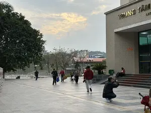 TRUNG TÂM LAO...。Quang cảnh tại một quảng trường lát gạch phía trước một tòa nhà. Nhiều người đang đi lại, một số mang theo túi và hành lý. Ở phía trước bên phải, một người đàn ông đang ngồi xổm chụp ảnh bằng điện thoại. Trên bậc thềm của tòa nhà, một người phụ nữ đeo khẩu trang đang ngồi xem điện thoại. Bầu trời có nhiều mây.。（日本語訳）。労働センター...。ある建物の前にある、タイル張りの広場の光景。多くの人々が行き交い、中にはカバンや荷物を持っている人もいる。右手前では、男性が一人しゃがんで携帯電話で写真を撮っている。建物の階段では、マスクをした女性が座って携帯電話を見ている。空には雲が多い。