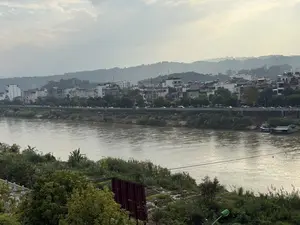 Bức ảnh chụp một khung cảnh ven sông vào một ngày nhiều mây. Một con sông rộng lớn với dòng nước màu nâu đục chảy ngang qua khung hình. Ở phía trước, bên bờ sông là cây cối và thảm thực vật xanh tươi, có một mảnh vườn rau nhỏ được trồng trên sườn dốc. Một người đang đội nón lá làm việc trong vườn. Ở bờ bên kia sông là một khu dân cư hoặc một thị trấn với nhiều tòa nhà san sát nhau. Một con đường chạy dọc bờ sông với nhiều ô tô đang đậu. Phía sau những ngôi nhà là những ngọn đồi cây cối rậm rạp, và xa hơn nữa là những dãy núi mờ ảo trong sương. Bầu trời u ám, bao phủ bởi những đám mây trắng xám, tạo nên một không gian yên tĩnh và có phần mờ ảo.。（日本語訳）。写真は曇りの日の川沿いの風景を写しています。濁った茶色の水が流れる大きな川が、画面を横切っています。手前の川岸には青々とした木々や草木があり、斜面には小さな野菜畑が作られています。ノンラーをかぶった一人の人が畑で作業をしています。川の対岸には、多くの建物が密集する住宅地または町があります。川岸に沿って道が走り、多くの車が駐車しています。空はどんよりとしており、白灰色の雲に覆われ、静かでやや幻想的な空間を作り出しています。