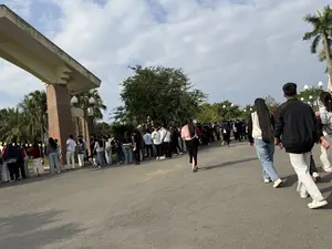 Một đám đông lớn các bạn trẻ đang tụ tập ngoài trời trên một con đường nhựa. Họ đang đứng thành từng nhóm, một số dường như đang xếp hàng gần một công trình cổng lớn có cột trụ bằng gạch. Khung cảnh xung quanh có nhiều cây xanh, bao gồm cả cây cọ, dưới bầu trời có mây. Mọi người mặc trang phục thường ngày như áo khoác, quần jean, và áo phông. Ở phía trước, một vài người đang đi bộ rời khỏi đám đông.。（日本語訳）。大勢の若者たちが、屋外のアスファルトの道に集まっています。彼らはグループになって立っており、何人かはレンガの柱がある大きな門の建造物の近くで列を作っているようです。周囲の景色には、ヤシの木を含む多くの緑の木々があり、曇り空が広がっています。皆、ジャケット、ジーンズ、Tシャツといった普段着を着ています。手前では、数人が群衆から離れて歩いています。