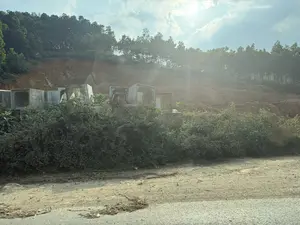 Bức ảnh chụp một khung cảnh ven đường, có thể là một công trường đang thi công. Ở phía trước là lề đường đất và các bụi cây xanh um tùm. Phía sau bụi cây, có nhiều khối cống hộp bê tông hình vuông được xếp chồng lên nhau. Nền phía sau là một vách đất đỏ lớn, trông như một ngọn đồi đã được san lấp. Trên đỉnh đồi là một hàng cây rừng rậm rạp. Bầu trời phía trên có mây và ánh nắng mặt trời chiếu sáng rực rỡ.。（日本語訳）。写真は道端の風景を写しており、おそらく工事中の現場です。手前には土の路肩と青々と茂る茂みがあります。茂みの後ろには、多くの四角いコンクリート製ボックスカルバートが積み重ねられています。背景は大きな赤土の崖で、造成された丘のように見えます。丘の上には、鬱蒼とした森の木々が並んでいます。上空には雲があり、太陽の光が明るく照らしています。