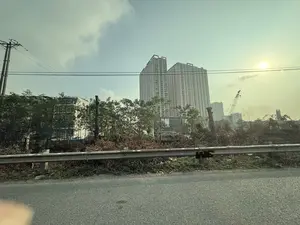 Bức ảnh chụp một cảnh quan đô thị, nhìn từ một con đường có lan can kim loại ở tiền cảnh. Phía sau lan can là một khu đất trống với cây xanh, cành cây khô và một cột điện. Ở phía xa, có hai tòa nhà chung cư cao tầng lớn và một số tòa nhà khác nhỏ hơn. Một chiếc cần cẩu xây dựng cũng hiện diện trong khung cảnh, cho thấy sự phát triển đô thị. Bầu trời có mây và hơi mờ, với ánh nắng mặt trời tỏa sáng ở phía trên bên phải.。（日本語訳）。写真は都市の景観を写したもので、前景に金属製の手すりがある道路から見ています。手すりの後ろには、緑の木々、枯れ枝、電柱がある空き地があります。遠くには、2つの大きな高層マンションと、その他いくつかの小さな建物があります。建設用クレーンも風景の中にあり、都市開発を示しています。空は曇っており、やや霞んでいて、右上から太陽の光が差し込んでいます。