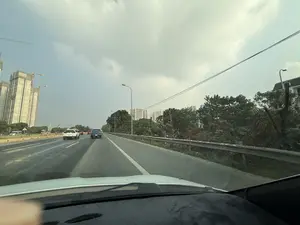 Bức ảnh được chụp từ góc nhìn bên trong một chiếc ô tô đang di chuyển trên đường cao tốc. Con đường có nhiều làn xe, phía trước có một vài chiếc ô tô khác đang chạy. Bên trái là một công trường xây dựng lớn với các tòa nhà cao tầng đang được thi công và nhiều cần cẩu. Bên phải đường là hàng cây xanh và lan can bảo vệ. Ở phía xa là đường chân trời của thành phố với nhiều tòa nhà chung cư. Bầu trời có nhiều mây.。（日本語訳）。写真は、高速道路を走行中の車内からの視点で撮影されたものです。道路は複数車線あり、前方には他の車が数台走行しています。左側には、建設中の高層ビルと多くのクレーンがある大きな工事現場があります。道路の右側には、緑の木々の列とガードレールがあります。遠くには、多くの集合住宅がある街のスカイラインがあります。空は雲が多いです。