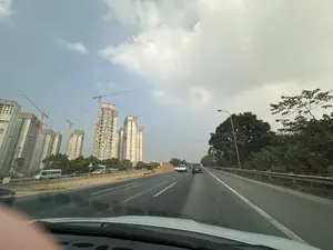 Quang cảnh được chụp từ bên trong một chiếc ô tô đang chạy trên đường. Phía trước là một con đường nhựa rộng rãi với nhiều làn xe. Bên trái đường là một công trường xây dựng quy mô lớn với nhiều tòa nhà chung cư cao tầng đang trong quá trình hoàn thiện, xung quanh có rất nhiều cần cẩu tháp. Bên phải đường là hàng cây xanh và lan can bảo vệ. Bầu trời có mây, một phần trong xanh và một phần u ám. Có một vài chiếc xe ô tô khác đang lưu thông trên đường.。（日本語訳）。道路を走行中の車内から撮影された光景です。前方には、複数の車線がある広いアスファルトの道路があります。道路の左側には、大規模な建設現場があり、多くの高層マンションが完成間近で、周りにはたくさんのタワークレーンがあります。道路の右側には、緑の並木とガードレールがあります。空は曇っており、一部は青く、一部はどんよりとしています。道路上には、他にも数台の車が走行しています。