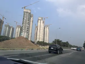 Khung cảnh một công trường xây dựng với nhiều tòa nhà cao tầng đang được thi công và các cần cẩu tháp. Trên các tòa nhà có tên công ty là 'NEWTECONS' và 'TEXO'. Phía trước là con đường có nhiều ô tô đang di chuyển. Một chiếc ô tô Mazda CX-8 màu đen mang biển số '19A 402.91'. Một chiếc xe khác màu trắng mang biển số '30H 178.04'.。（日本語訳）。多くの高層ビルが建設中で、タワークレーンがある建設現場の光景。ビルには「NEWTECONS」と「TEXO」という会社名がある。手前には多くの車が走行している道路がある。ナンバープレート「19A 402.91」を付けた黒いマツダCX-8が1台。ナンバープレート「30H 178.04」を付けた別の白い車が1台。