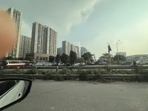 Bức ảnh chụp khung cảnh đô thị với nhiều tòa nhà chung cư cao tầng ở phía sau. Bầu trời có mây. Phía trước các tòa nhà là một con đường và bãi đậu xe với nhiều ô tô và một chiếc xe buýt màu đỏ. Có cây xanh và cột đèn đường. Ở bên phải, có một người đang đi xe máy. Tiền cảnh là một phần đường nhựa, gương chiếu hậu của ô tô và dải phân cách bằng kim loại có cây cối mọc um tùm.。（日本語訳）。背景に多くの高層マンションが建ち並ぶ都市の風景を写した写真です。空は曇っています。建物の手前には道路と駐車場があり、多くの車と1台の赤いバスが停まっています。緑の木々や街灯があります。右側には、バイクに乗っている人が一人います。